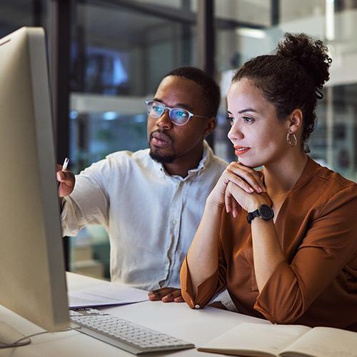 A man is pointing to his computer screen while talking to a coworker.