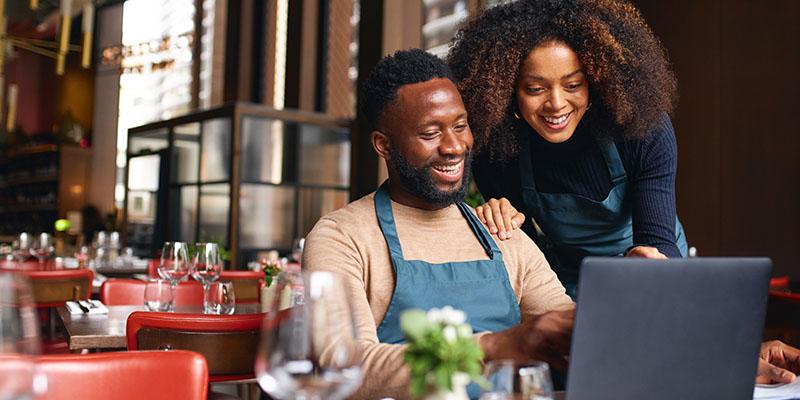 A woman and man smiling at a laptop in a restaurant setting.