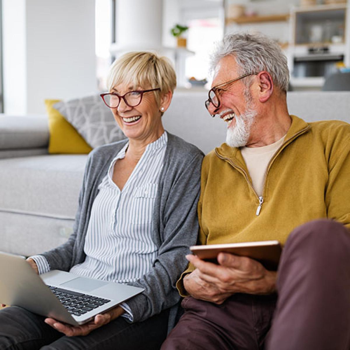 A couple sits on the floor near their couch holding a laptop and tablet while smiling.