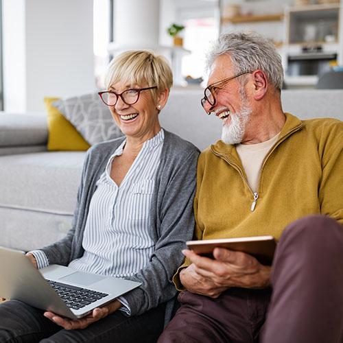 A couple sits on the floor near their couch holding a laptop and tablet while smiling.